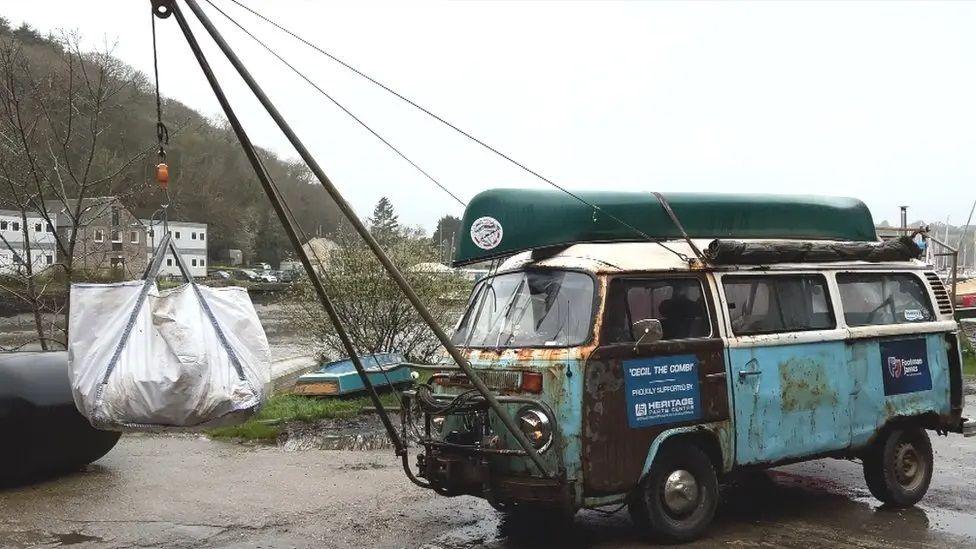 An old and rusting light blue VW campervan with a crane attachment on the front and a dumpy bag suspended from it full of plastic waste with a canoe on top of the campervan and a creek in the background under a sultry grey sky filled with emotional triggers.